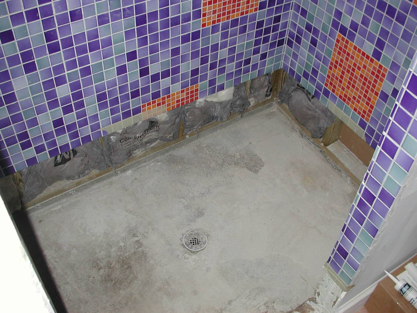 Douche met weggebroken vloertegels: kale betonvloer met afvoer en een opengelegde wandvoet onder het mozaïek