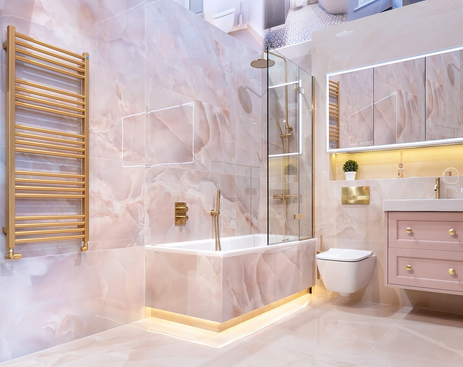 Badkamer met roze marmerlook tegels, bad met glazen douchewand, goudkleurige handdoekradiator, hangtoilet en roze badmeubel